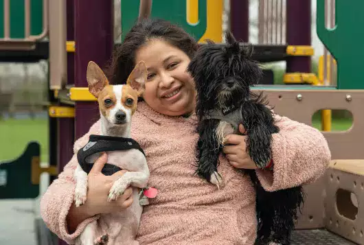 A woman sits at a playground smiling with a small dog in each arm